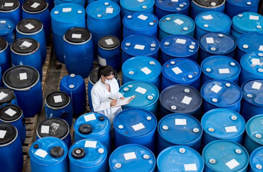 Woman in a lab coat inspecting blue barrels in a storage area with labels on them.